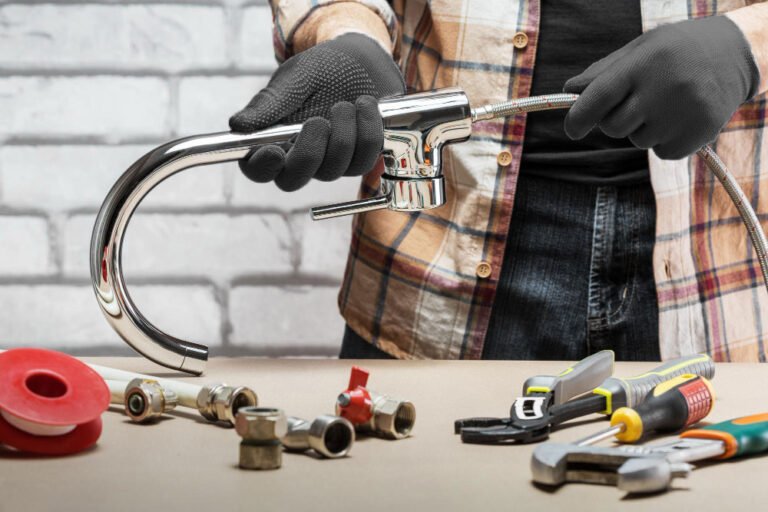 Plombier tenant un robinet chromé avec flexible, entouré d’outils et de pièces détachées.
