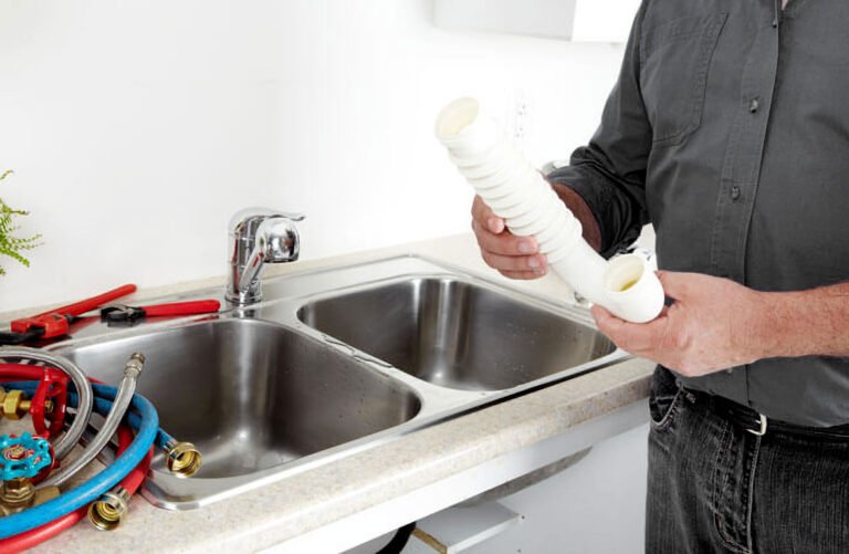 Plombier tenant un tuyau en PVC blanc pour remplacer un ancien raccord sous un évier double.