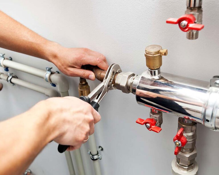 Plombier utilisant une pince pour serrer un raccord sur un chauffe-eau avec tuyaux en PVC et vannes rouges.
